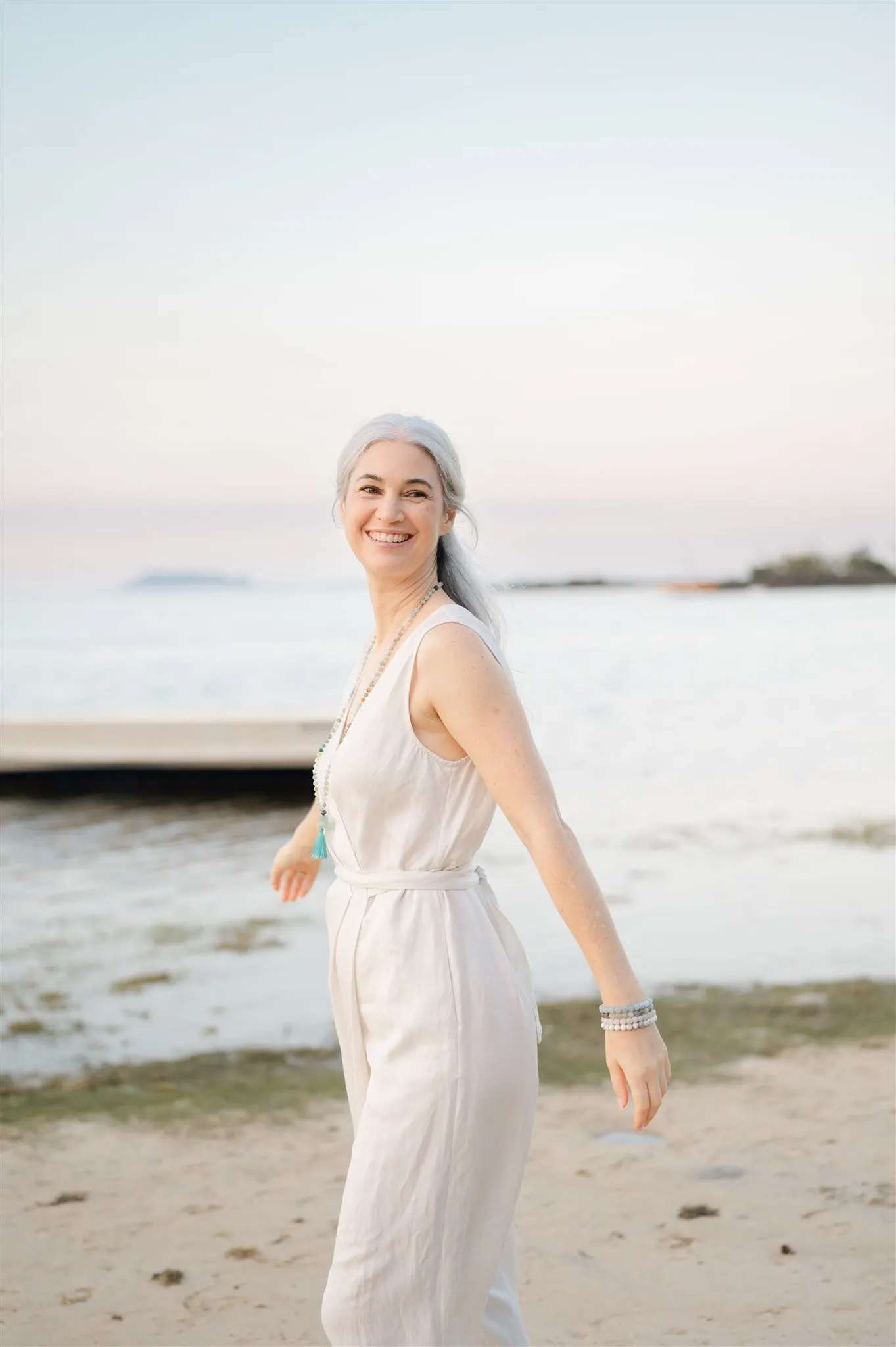 Stephanie turning on the beach at dusk in a pale linen jumpsuit, arms open